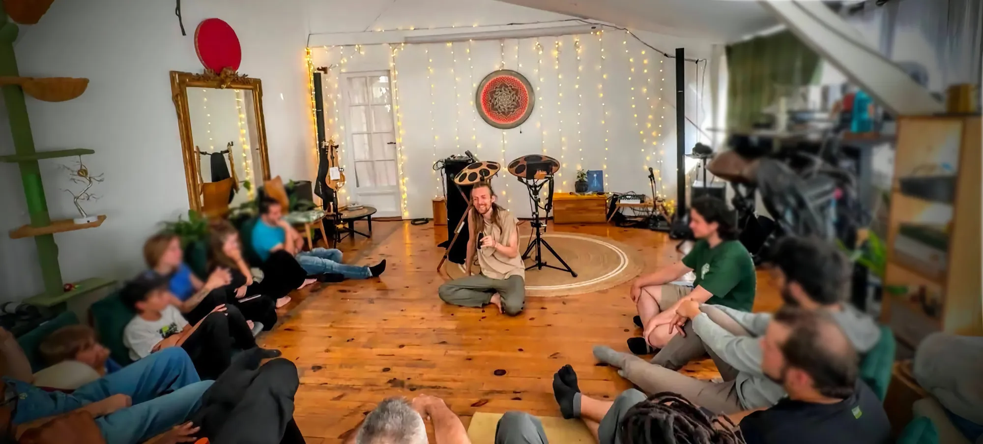 Vue d’ensemble d’une rencontre au Nid : David Lesage assis au centre du cercle, handpans sur pieds derrière lui, le public assis à même le parquet, murs tendus de guirlandes lumineuses et rosace suspendue.