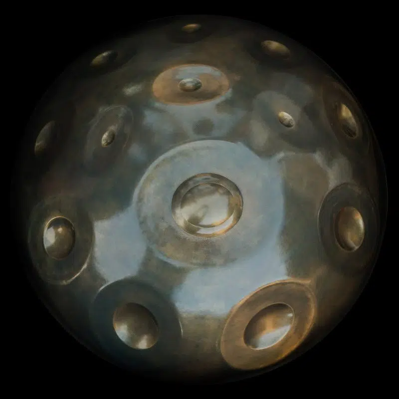 Handpan Yishama A Minor 17 visto desde arriba sobre fondo negro: superficie gris azulada moteada con reflejos bronce, el ding en el centro y una quincena de zonas de notas alrededor.
