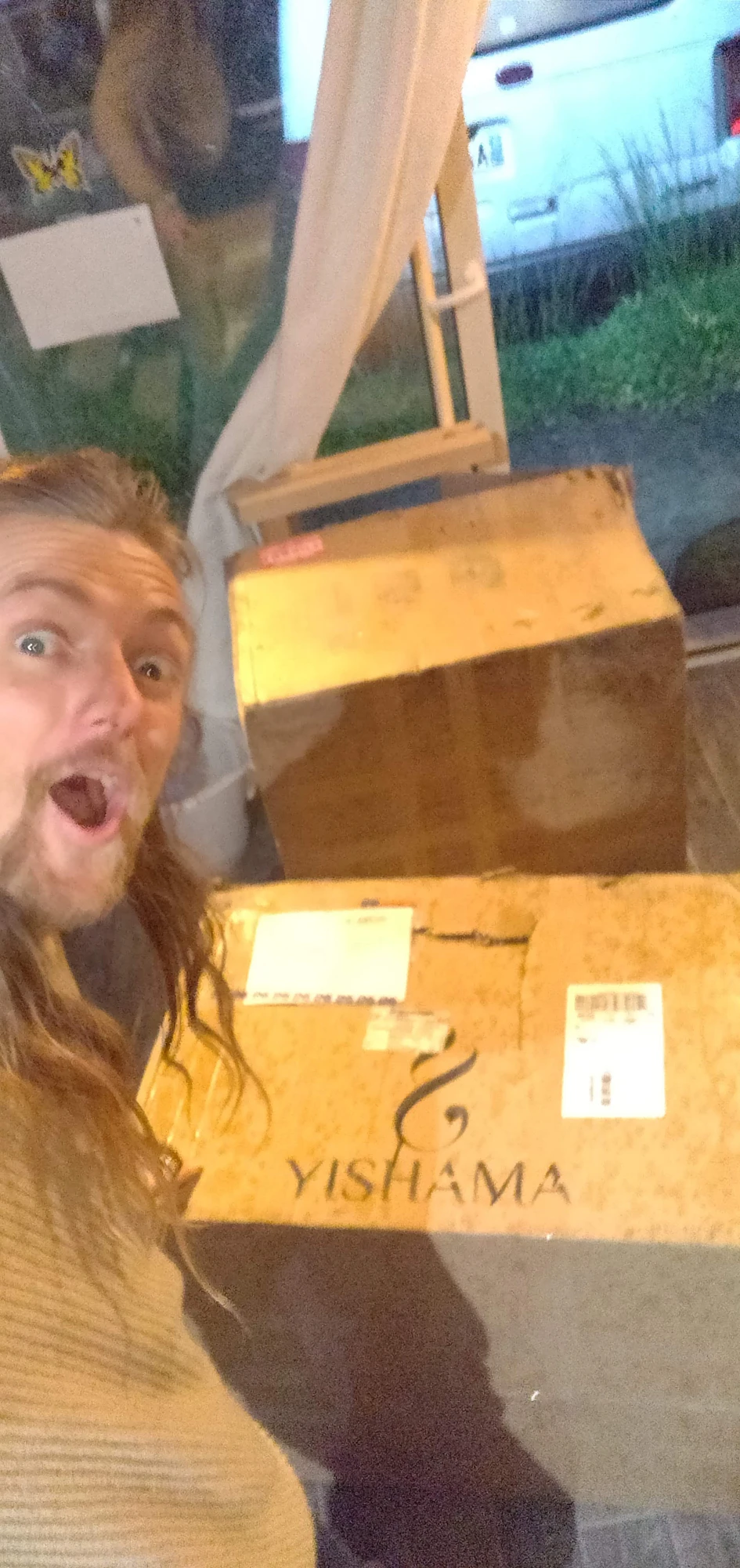 Una foto borrosa tomada al atardecer: David Lesage en primer plano extremo, con la boca y los ojos muy abiertos. Detrás de él, dos grandes cajas de envío están apiladas; la de abajo lleva el logotipo y la palabra YISHAMA en letras grandes, junto con sus etiquetas de envío. Al fondo, tras la puerta de cristal, el camión de reparto blanco sigue aparcado frente a la casa.