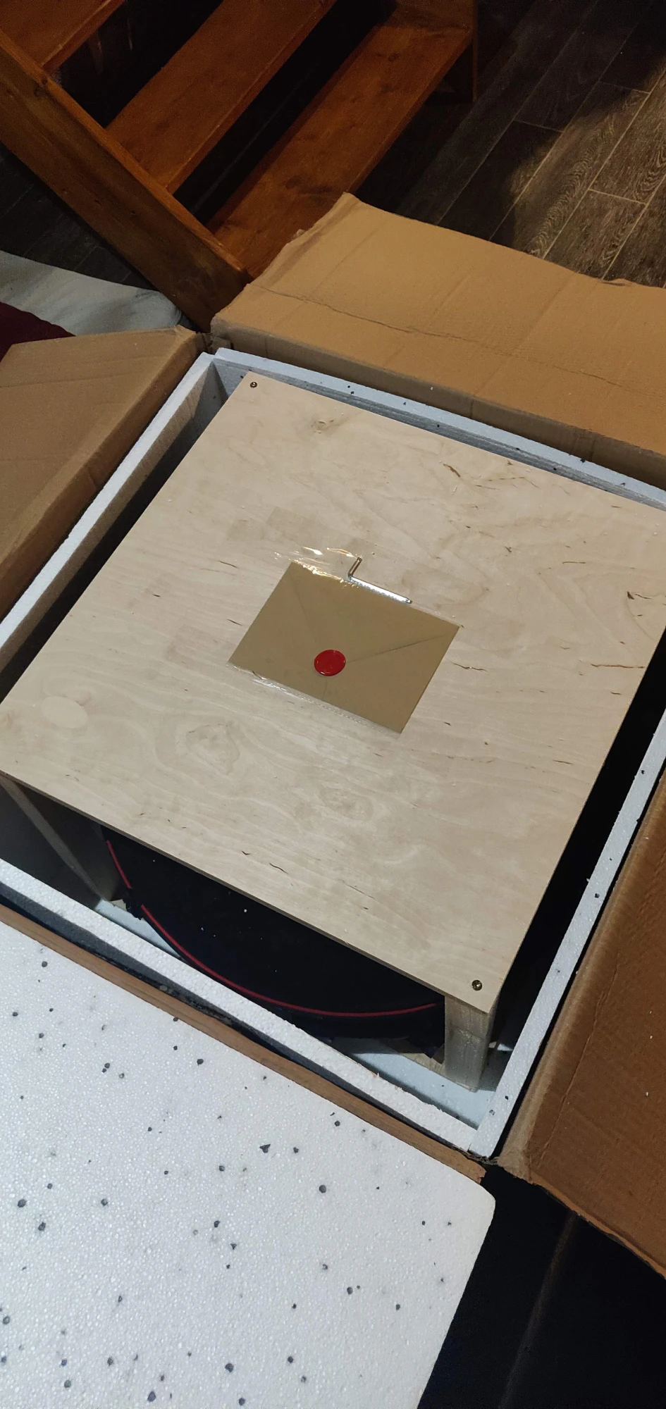 Vista aérea de la caja abierta: acolchado de poliestireno blanco y, en el interior, una caja de madera contrachapada ligera asegurada con tornillos visibles en las esquinas. Sobre la tapa, dentro de una funda protectora de plástico, hay un sobre de papel kraft sellado con cera roja. A través de la abertura bajo la tapa, se ve la cubierta negra con ribete rojo.