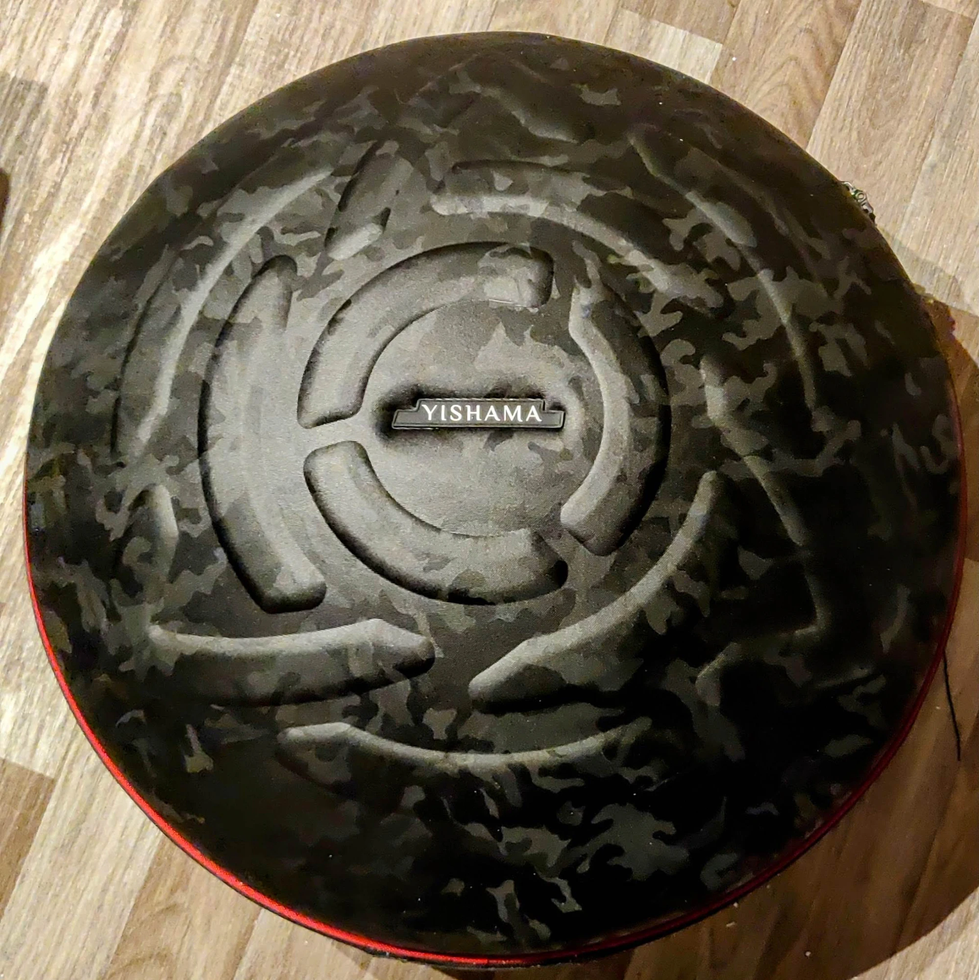 La cubierta rígida del handpan vista desde arriba, colocada sobre un suelo de parqué claro: tela de camuflaje negro mate, espiral ancha acolchada en relieve, placa rectangular con la palabra YISHAMA en el centro, ribete rojo en el borde y cremallera alrededor del perímetro.