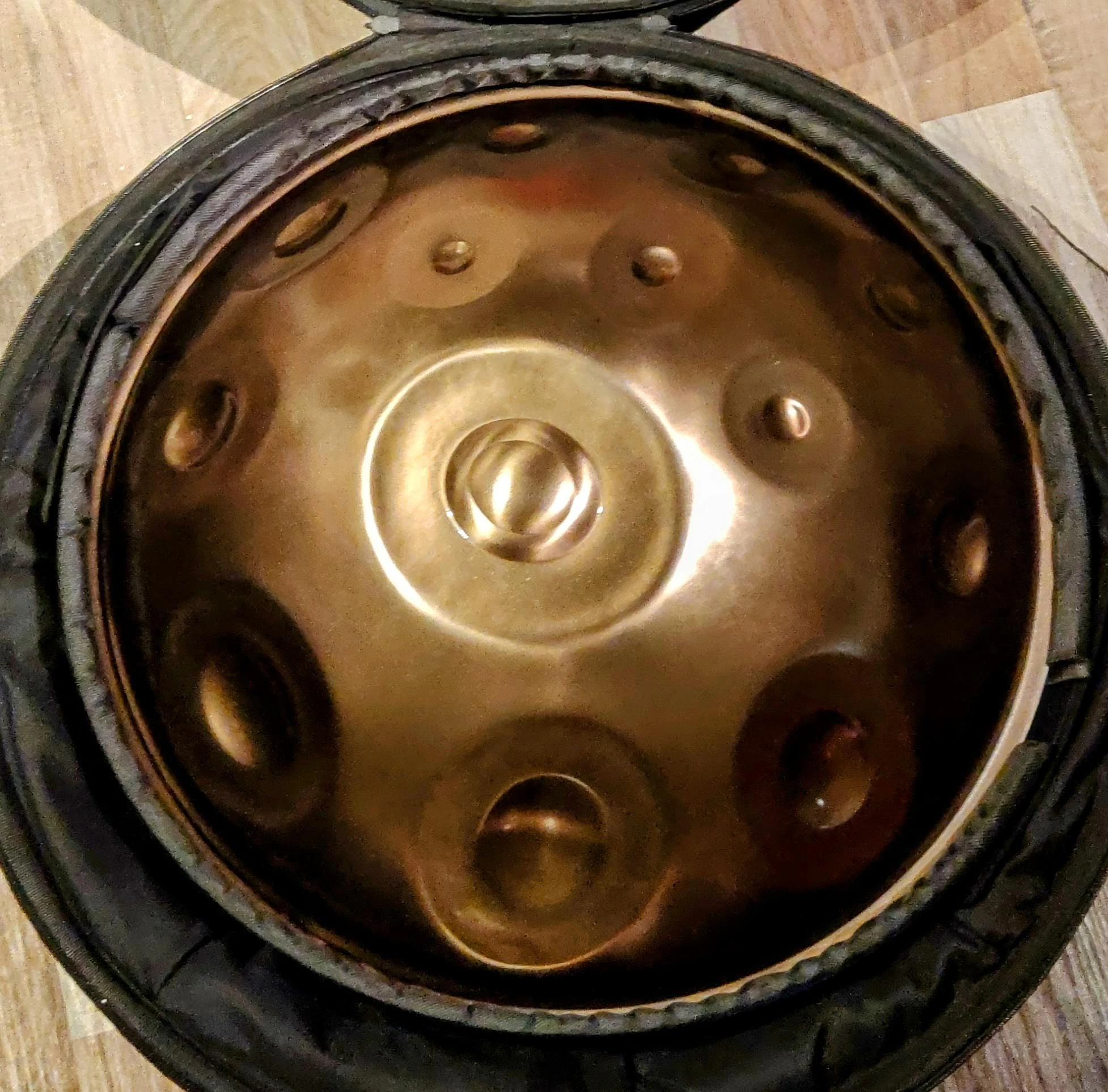 La vitrina abierta deja ver el handpan, cuyo metal refleja una luz cálida, dorada y bronceada. En el centro se encuentra el ding y sus anillos concéntricos; a su alrededor, las zonas de notas, en huecos y cúpulas.