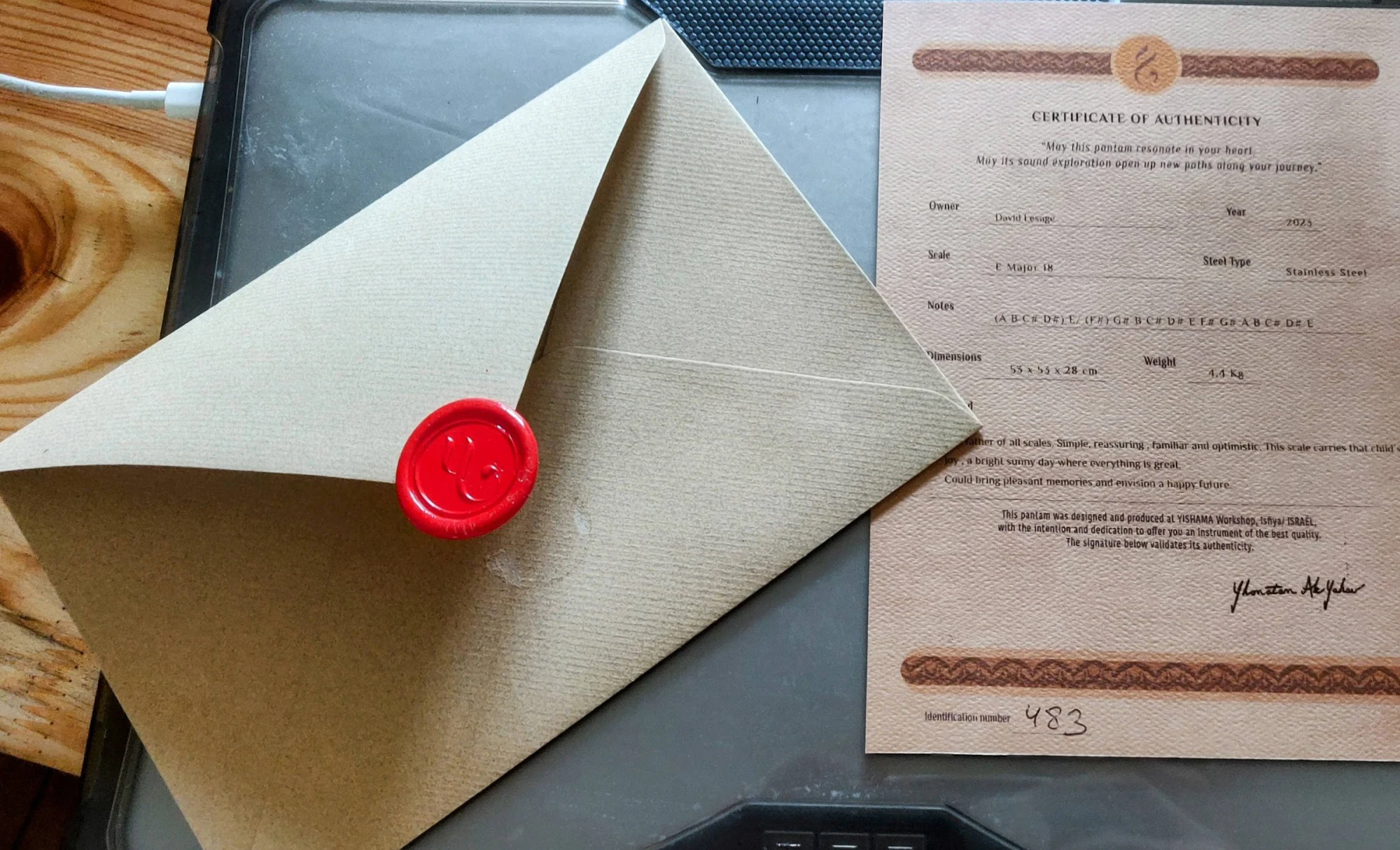 Sobre una superficie de trabajo de madera: el sobre de papel kraft, ahora abierto, con su sello de cera roja que lleva un monograma, colocado junto al certificado de autenticidad impreso en papel texturizado.