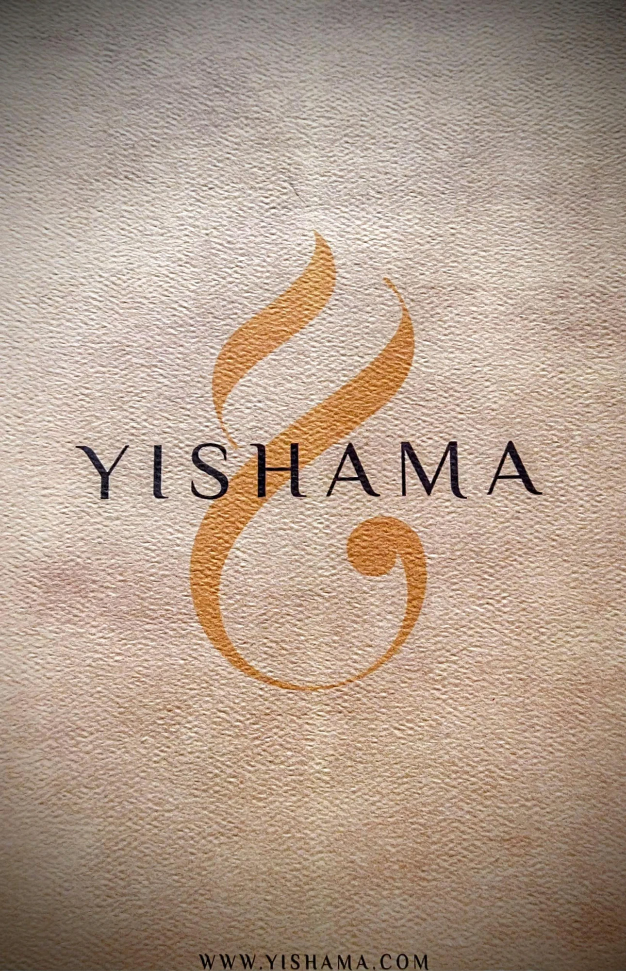 El reverso del certificado: el logotipo Yishama —un pergamino dorado— sobre la palabra YISHAMA en mayúsculas, en papel texturizado, con la dirección WWW.YISHAMA.COM en la parte inferior de la página.