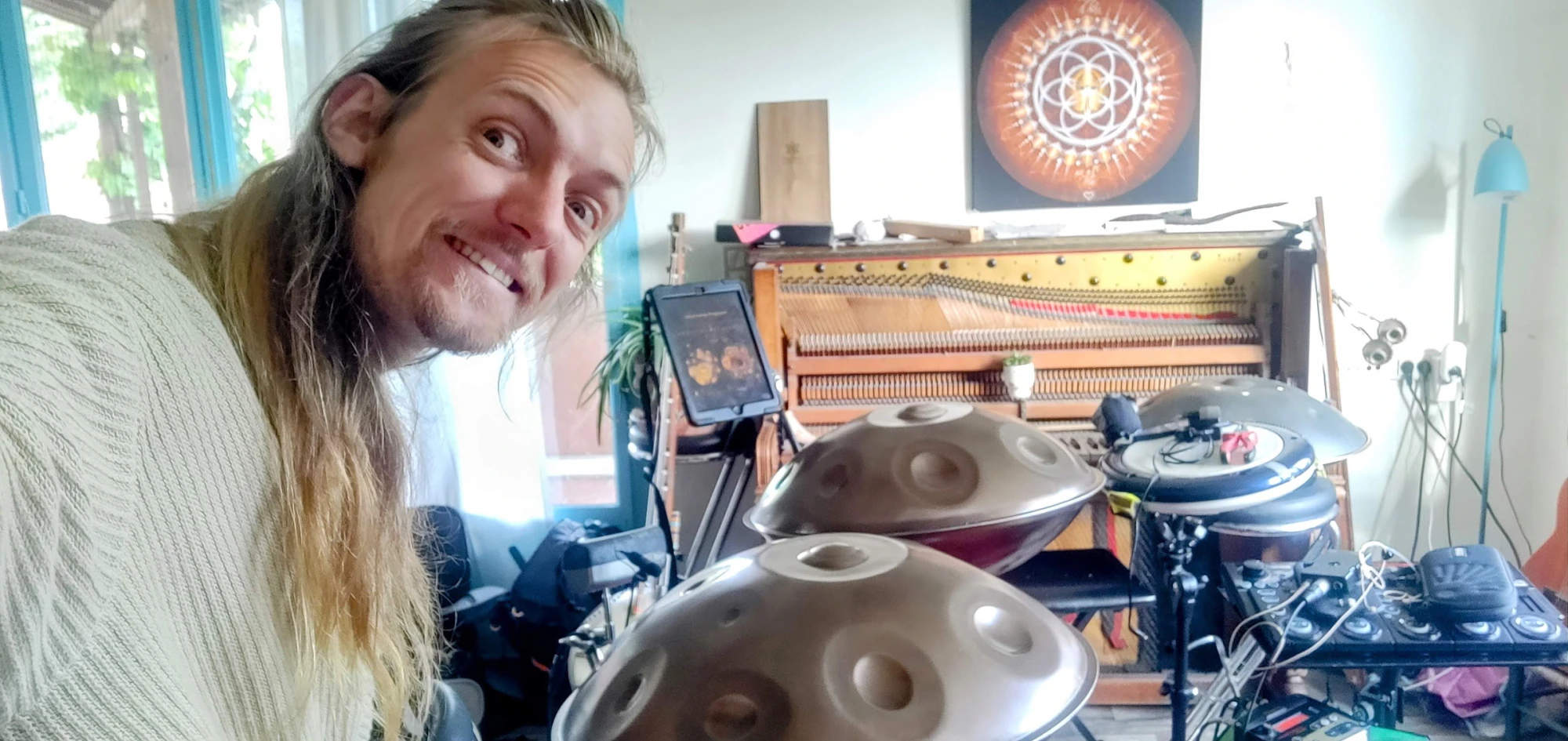 David Lesage sonriendo, con la cabeza inclinada, a plena luz del día. Delante de él, sus dos handpans Yishama de metal gris antracita apoyados sobre sus bases; detrás, una tableta fijada a un brazo articulado, el marco de un piano vertical abierto que deja ver las cuerdas y la mecánica, una pintura que representa una Flor de la Vida naranja, pads electrónicos y un pedalier.